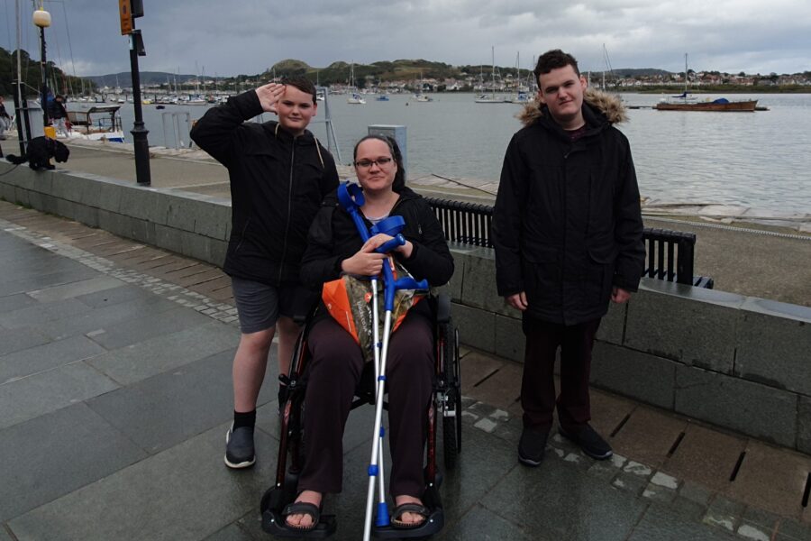 In front of a wide river, a woman in a wheelchair smiles at the camera, with two young men either side of her. One of the boys is saluting the camera.