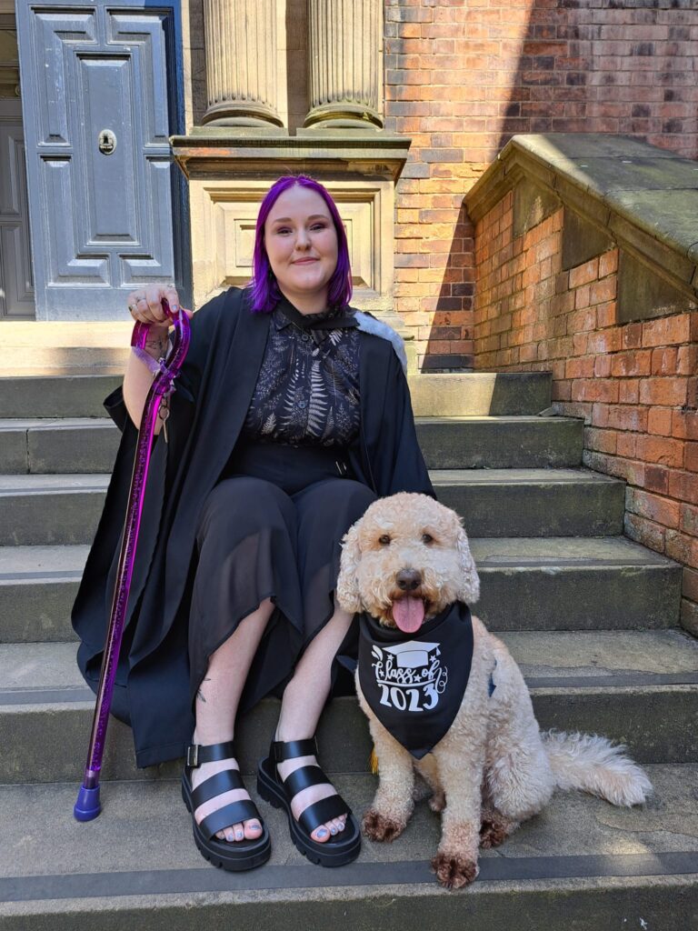 Lauren Gilbert, a white person with purple hair, sitting on the front steps of a building with their dog, smiling and holding a walking stick.