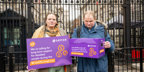 2 people stand in from of the railings on Parliament Square. They are holding a poster and box of petitions asking for the government to consider the additional costs of living for disabled people.