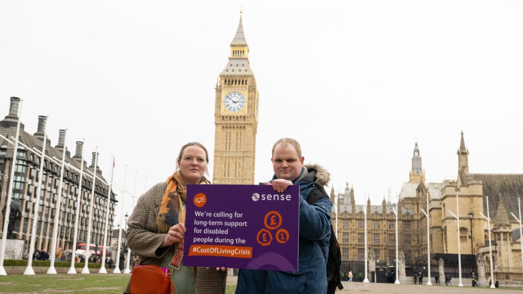 A man and woman standing in Westminster hold a placard. It reads 'We're calling for long term support for disabled people during the cost of living crisis'.