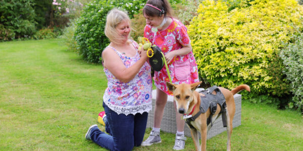 A young girl stands in a garden with her dog while her mum kneels beside her