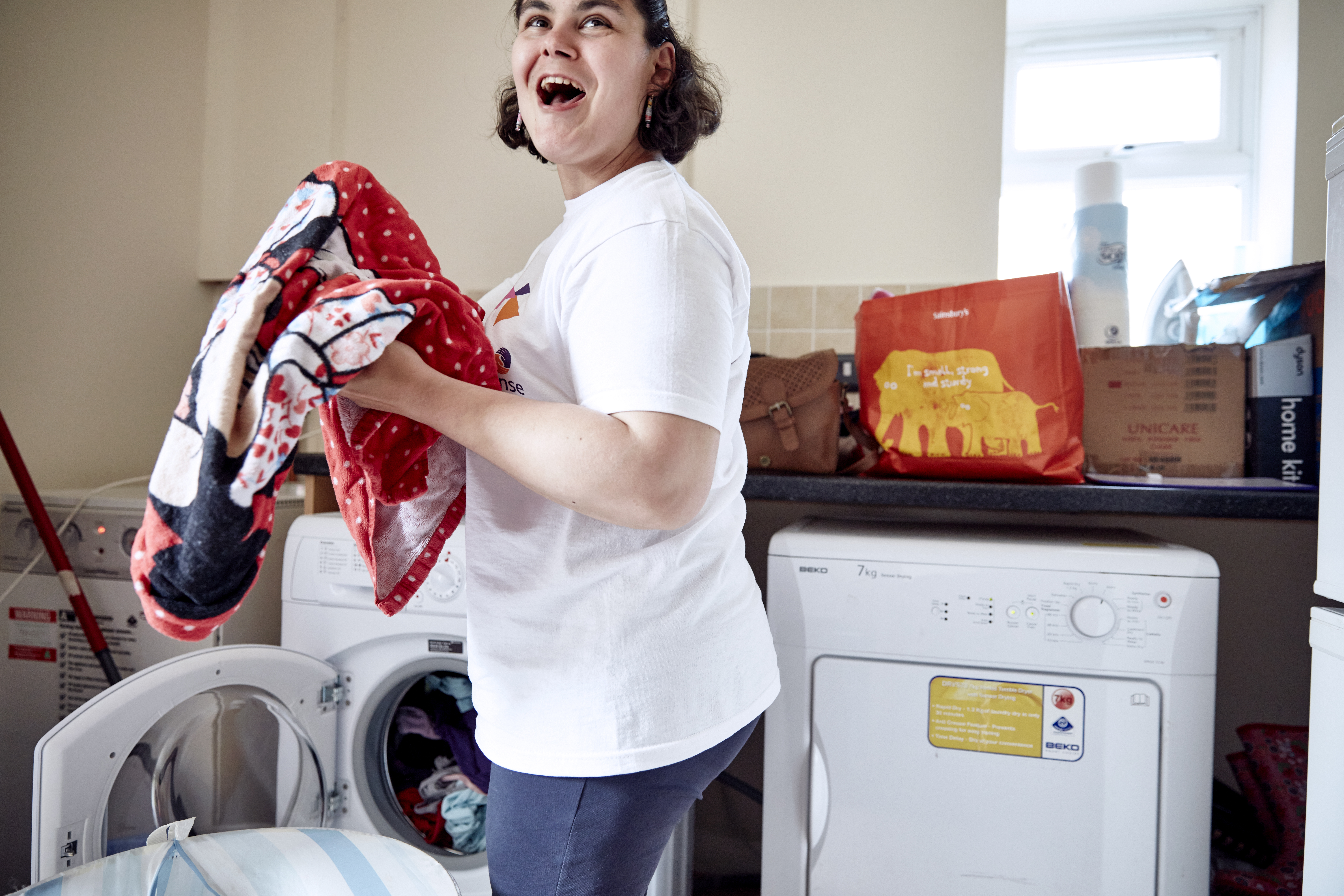 A woman with dark curly hair puts laundry in the washing machine while laughing.