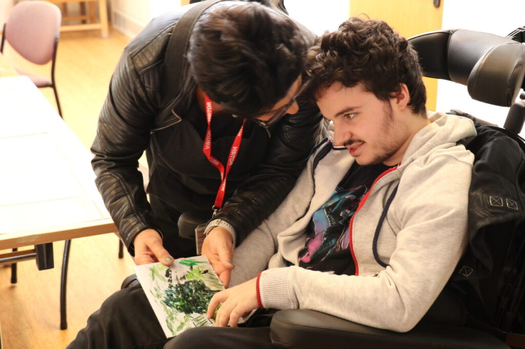 A man leans over to chat to a boy in a wheelchair. His painted artwork sits in his lap.