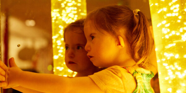 A young girl stands next to a mirror so we see her and her reflection. Behind her is a sensory bubble tube, full of bubbles and illuminated in orange light.