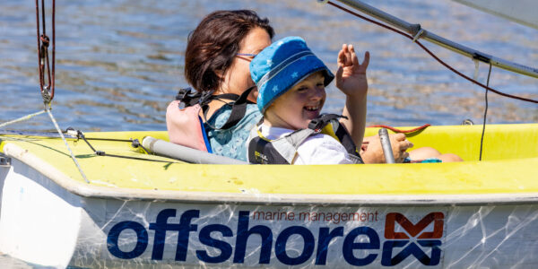 A young boy is in a yellow and white sailing boat with the words 'offshore' in blue text on the side. He is wearing a blue bucket hat and waving an arm in the air.
