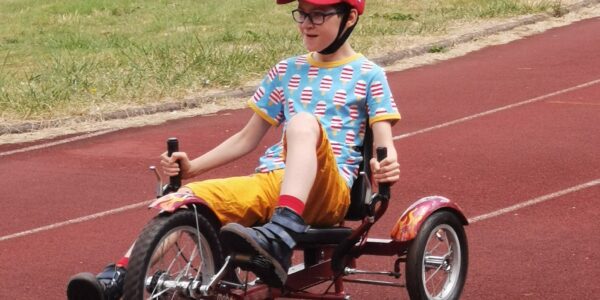 A young boy riding an adapted bike around a red race track. He is wearing a blue patterned tshirt, orange shorts and a red cap.