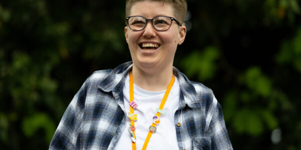 A woman with short croped brown hair wearing glasses laughs to the camera. She is wearing a white tshirt with a blue, black and white checked shirt over the top and an orange lanyard with lots of pin badges attached.