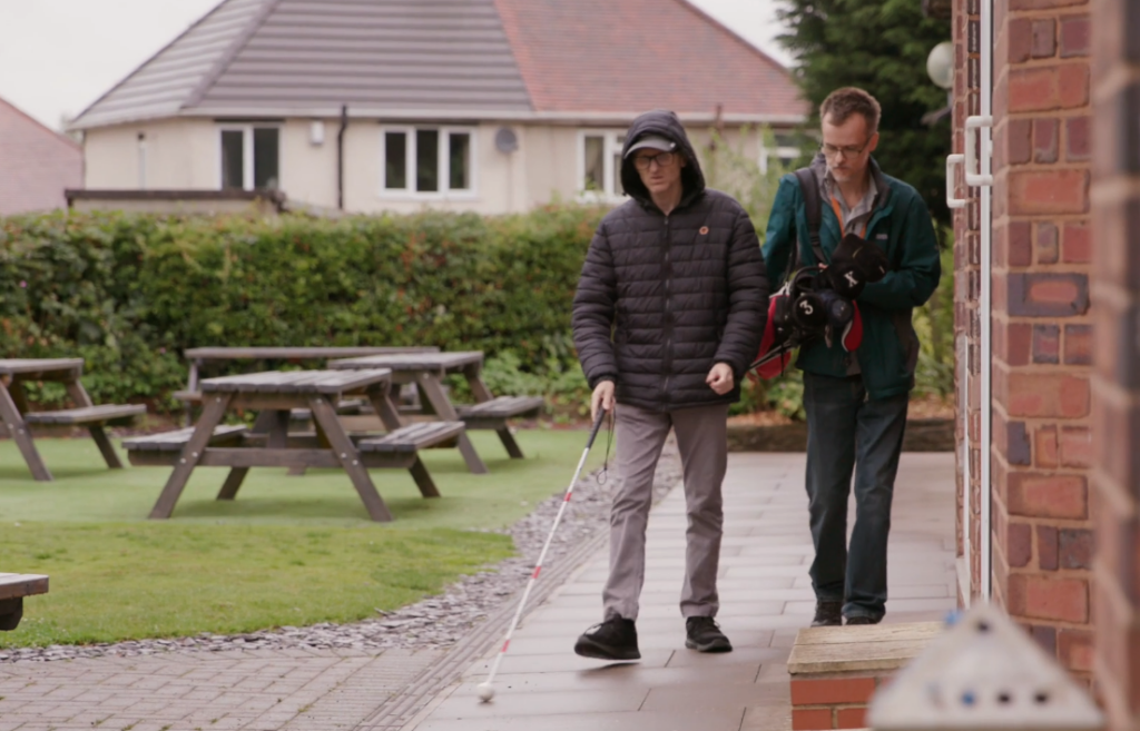 Simon walks towards the camera using a white cane to identify the path. Behind him, another man carries Simon's golf clubs.