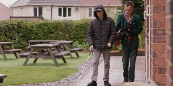 A man uses a cane to walk assisted by another man holding his free arm