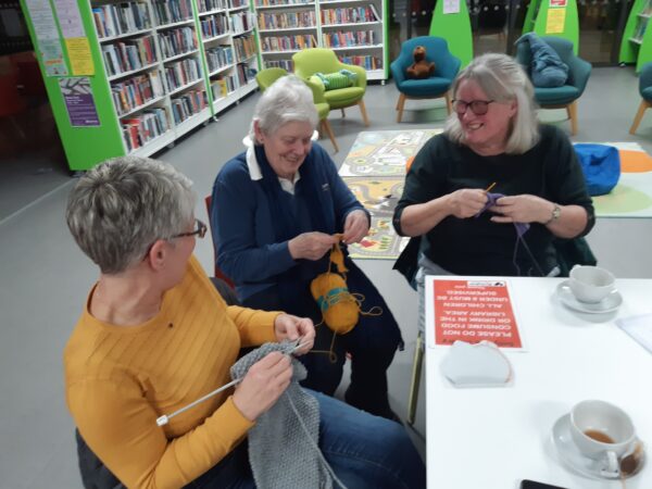 3 women, laughing whilst knitting at a table.