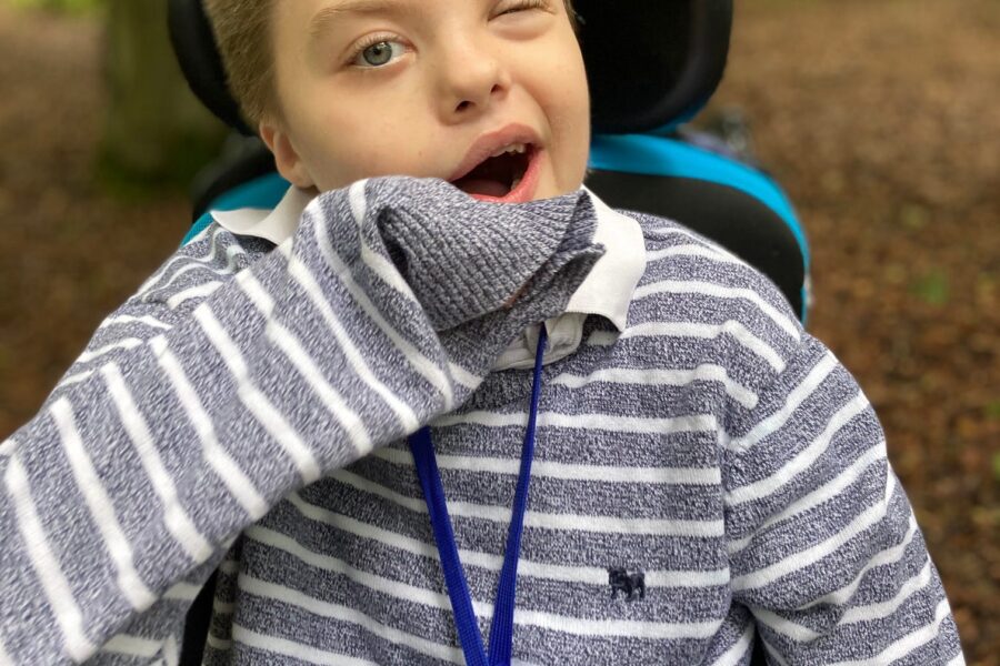 A young boy in a powerchair wearing a grey and white striped jumper. He also has a necklace around his neck with a blue pendant on. Behind him is a woodland.