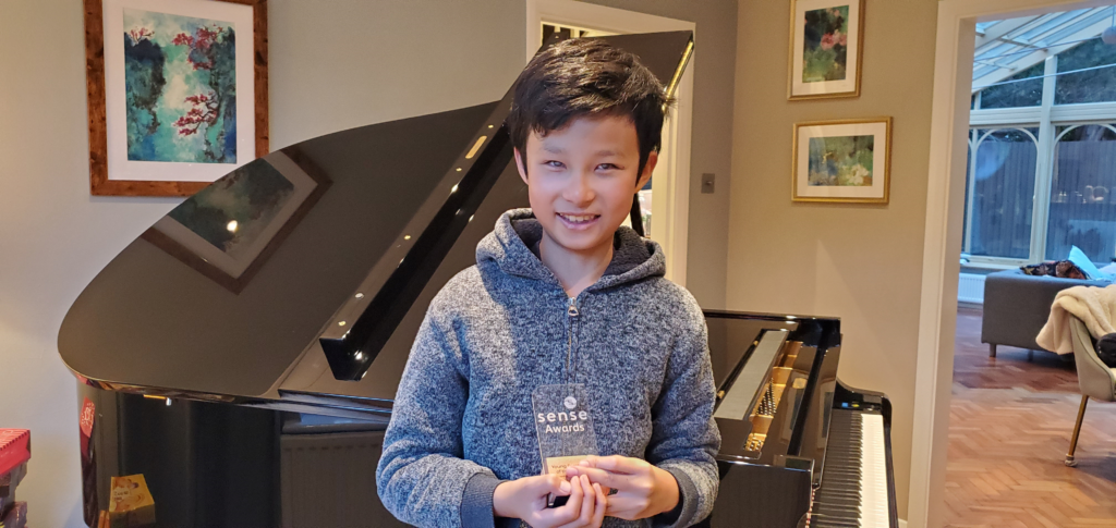 A young boy in a grey jumper holding a trophy in front of a piano