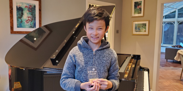 A young boy in a grey jumper holding a trophy in front of a piano