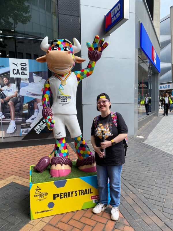 A woman weaing glasses, a cap, a black tshirt and blue jeans is standing next to a statue of a multicolored bull. The bull is wearing a white top that says 'Birmingham 2022'.