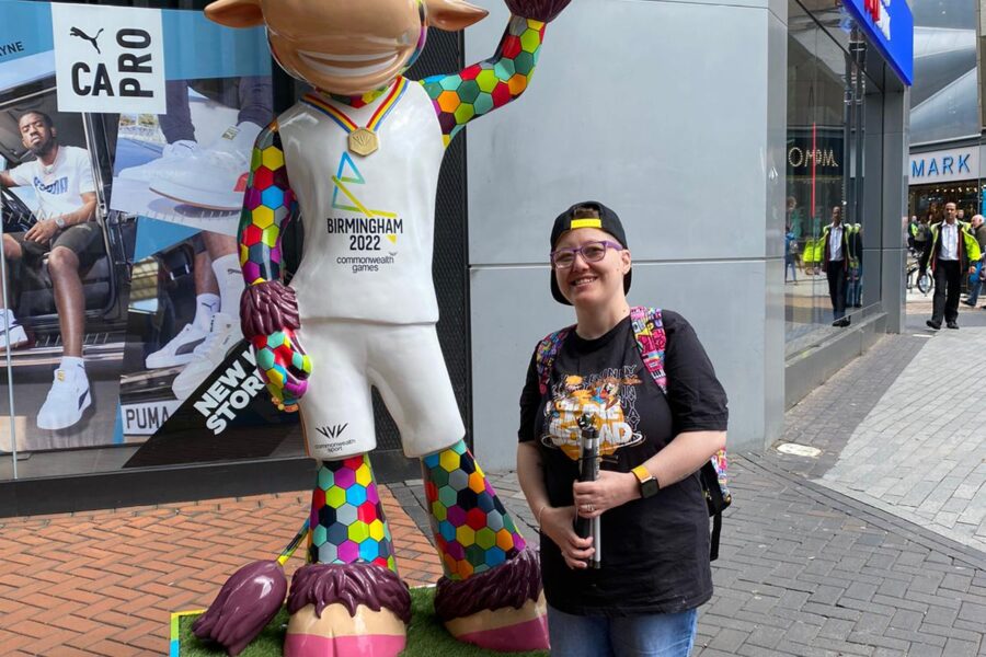 A woman weaing glasses, a cap, a black tshirt and blue jeans is standing next to a statue of a multicolored bull. The bull is wearing a white top that says 'Birmingham 2022'.