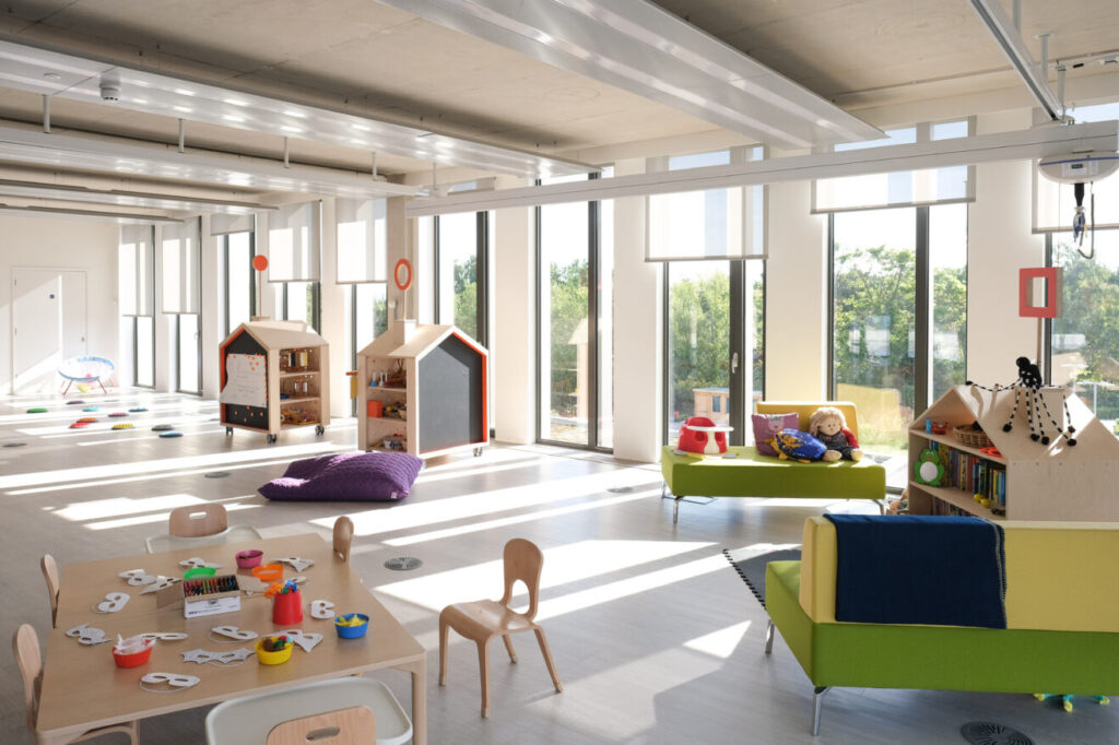 A large room with big windows and sunlight streaming through. It is set up for children with toys, books, sofas, and different play spaces.