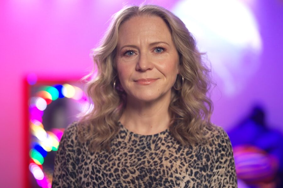 Kellie Bright smiles at the camera in front of a pink and purple background