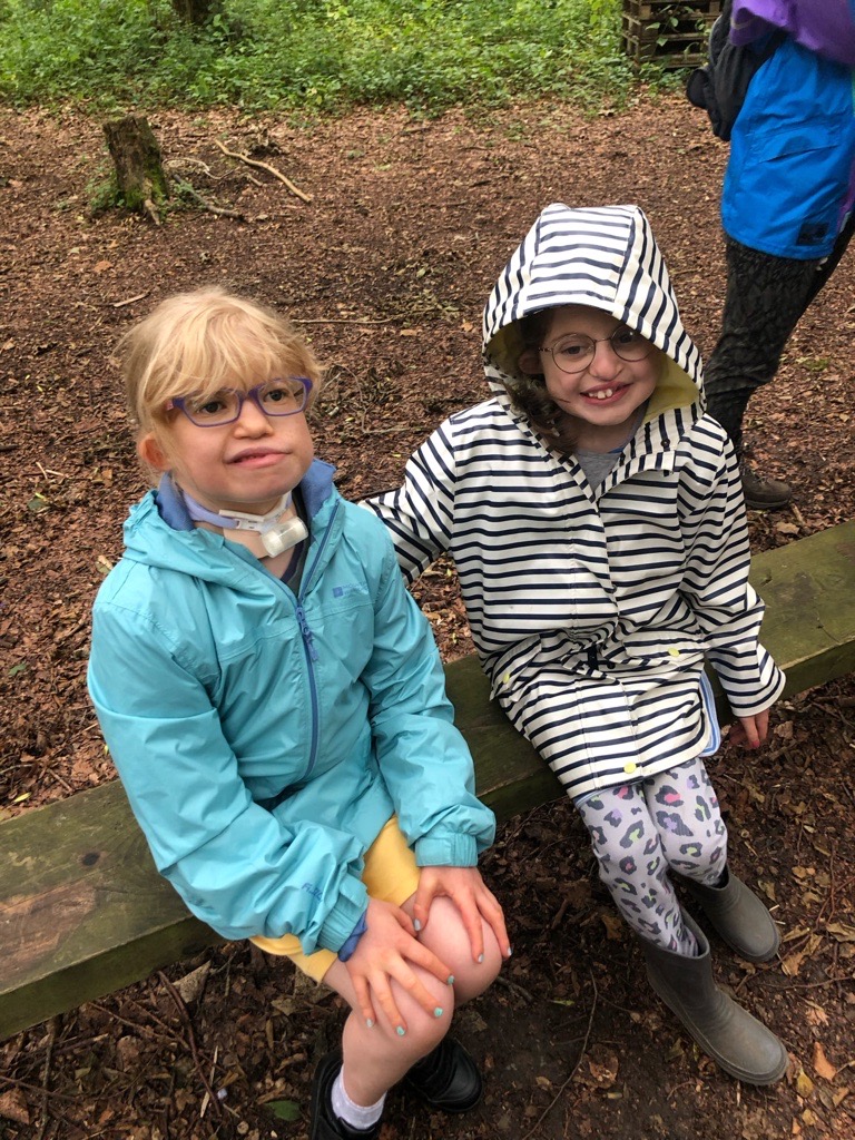 Friends Orla and Bethany wearing their raincoats and smiling while sitting on a bench in the forest.