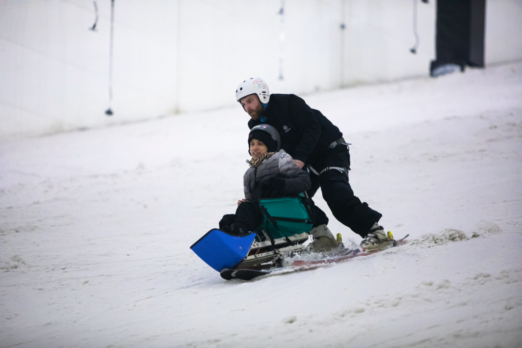 A ski instructor in a black ski suit and white helmet skis down a slope with a young man sat in a sit ski