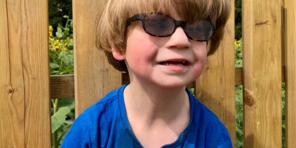 A boy in a blue t-shirt witha a huge mop of blond hair looks tot he right of the camera. He wears dark glasses in the sunshine.