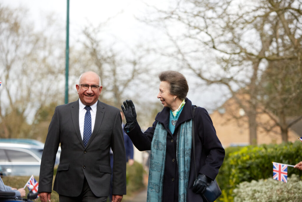 Her Royal Highness The Princess Royal is walking next to a man in glasses and a suit. She waves and smiles to people on her right as she walks
