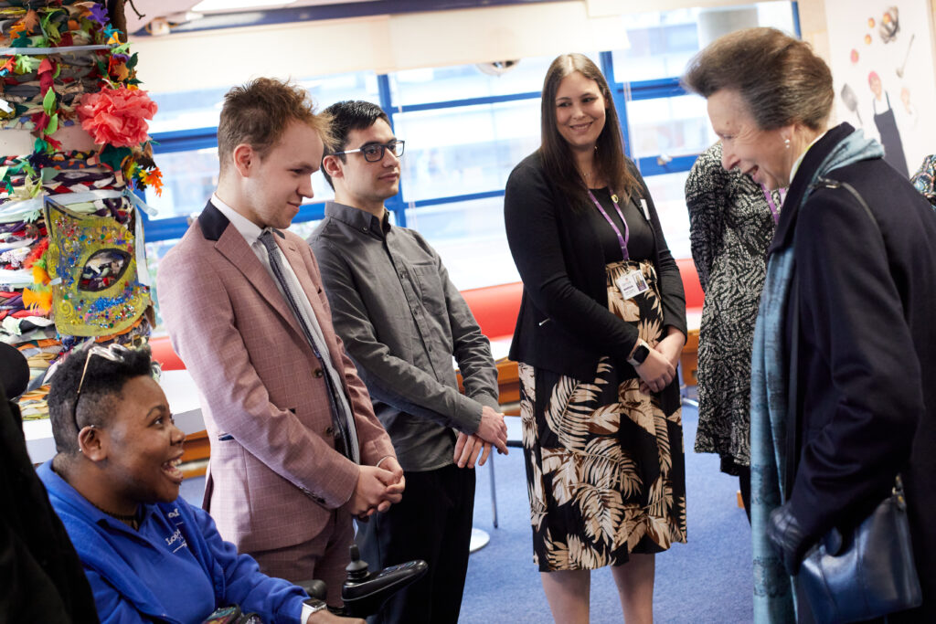 Her Royal Highness The Princess Royal greeting people in a room. There is a young man in a wheelchair, next to him is a man standing in a pink suit, a man standing in a grey shirt and a lady standing wearing a patterned black dress.