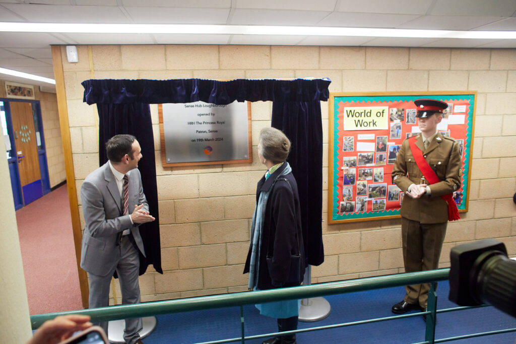 Her Royal Highness The Princess Royal stands looking at a commemorative plaque on a wall. The plaque is surrounded by a blue curtain. Richard Kramer, Sense CEO stands next to her in a grey suit. To her right is a man in a green military uniform.