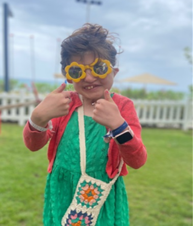 Orla, a young girl in a green dress and yellow, flower-shaped sunglasses, grins and gives a double thumbs up to the camera.