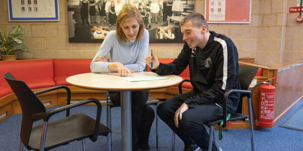 Two people sitting at a table. They are looking at a piece of paper on the table in front of them and talking about it.