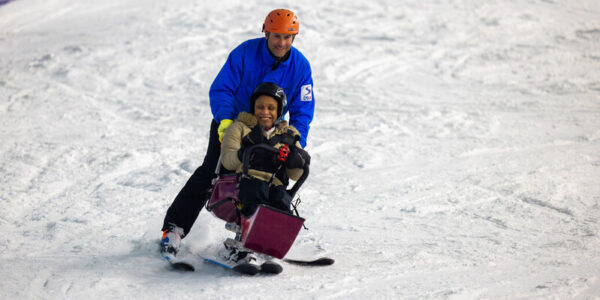 A woman on a sit ski is guided down a snowy slope by her instructor, who's skiing close behind her.