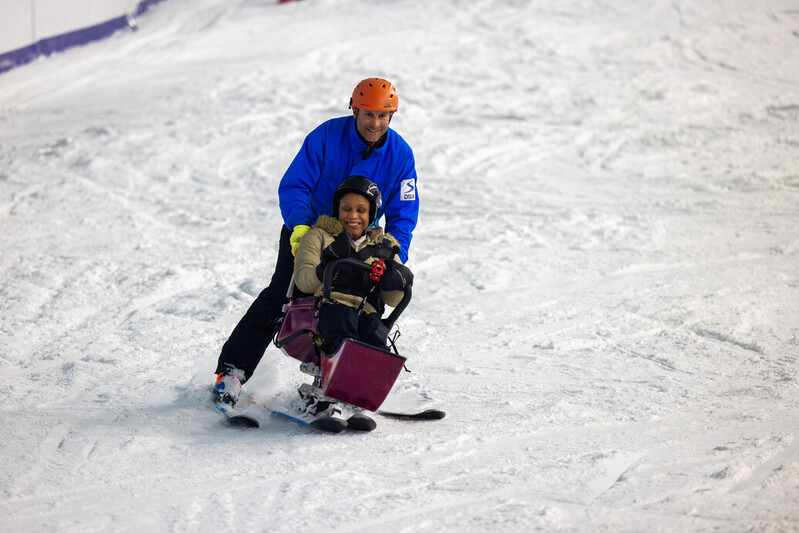 A man in blue coat pushes a smiling woman in a sit ski down a snowy slope.