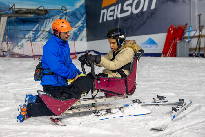 Hazel sits in a chair connected to two skis. Her instructor James kneels beside Hazel.