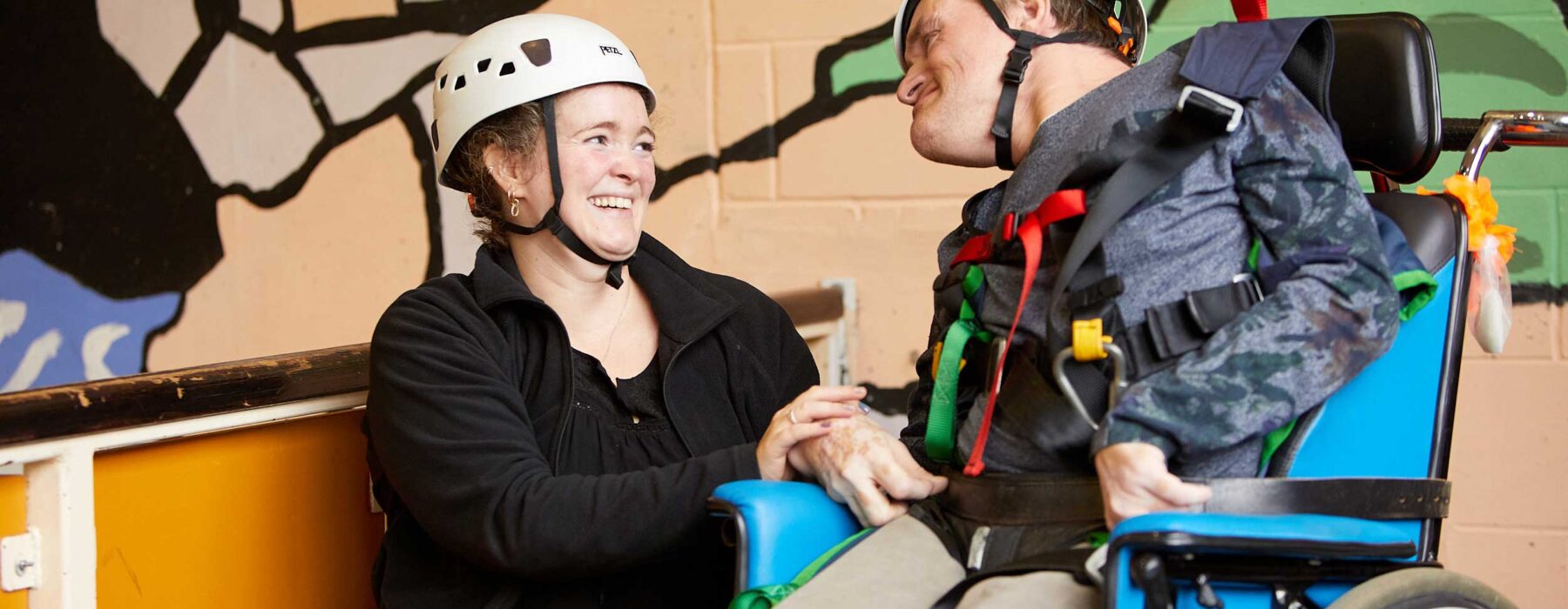 A woman kneels next to a man in a wheelchair. Both are wearing hard hats for climbing.