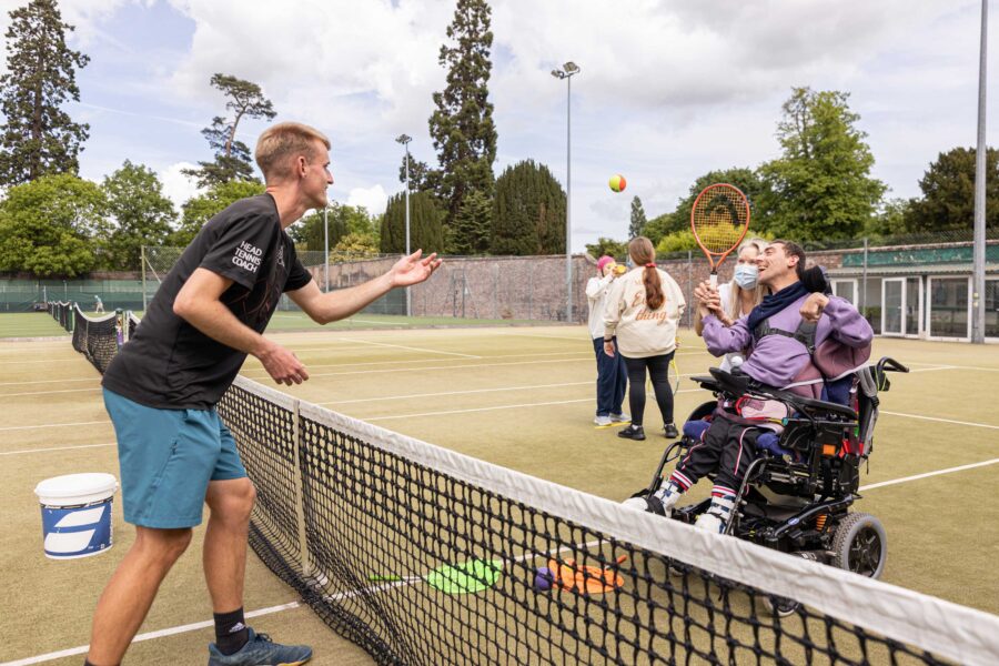 A man on the left of a tennis net throws a ball to a delighted man in a wheelchair on the other side. the man in the wheelchair is holding a racquet.