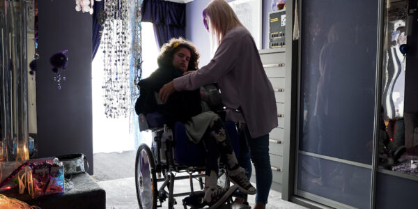 Yvette, a white woman with blonde and purple hair, putting a jacket on her daughter Rosey, a young woman with brown hair in a wheelchair.