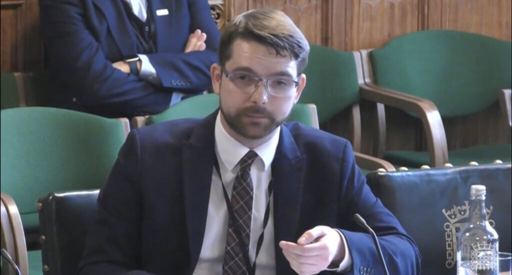Evan wearing a suit sat at a table in parliament. His hand is raised as he is making a point when he speaks.