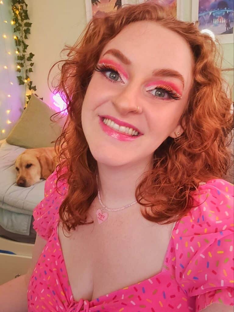 A woman with red hair and brightly coloured eye make up smiles at the camera, wearing a bright pink top with multicoloured dots on it. Behind her, there's a golden retriever dog resting its head.