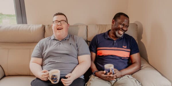 Jamie sharing a laugh with a support worker as they both drink a cup of tea on the sofa.