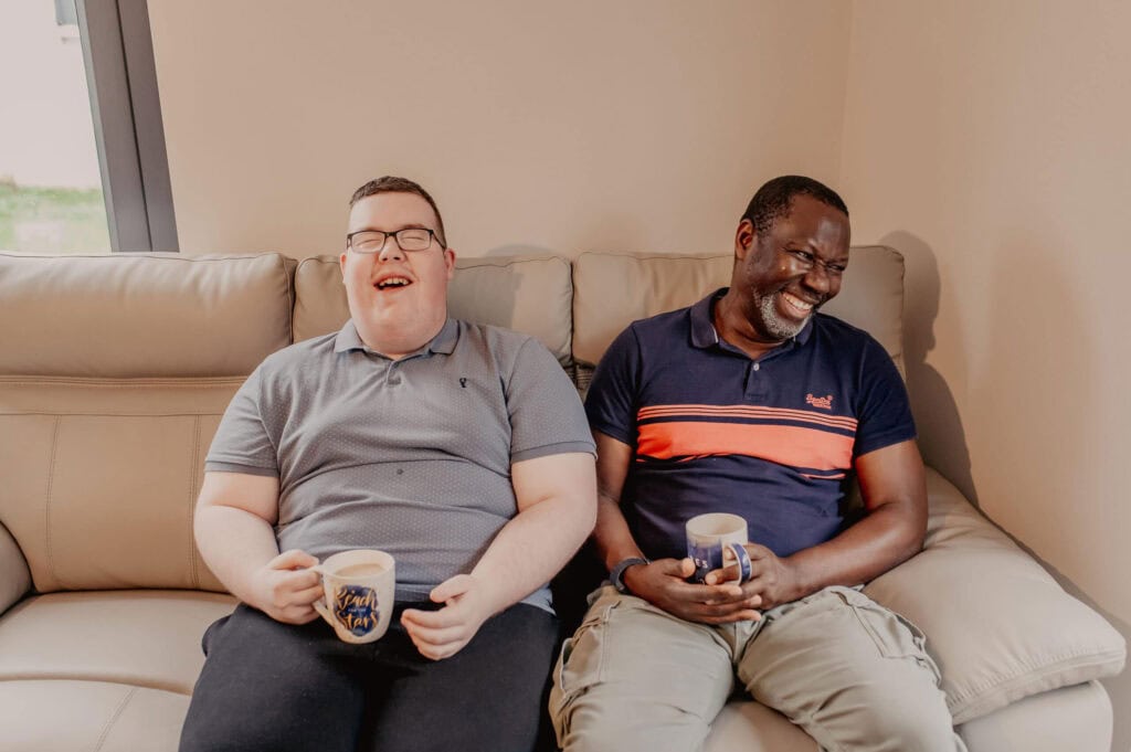 Two men are sitting next to each other laughing to each other. They're both holding cups of tea in their hands.