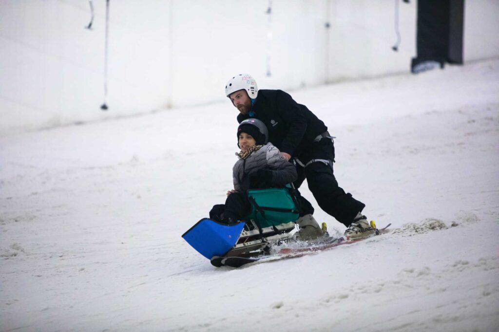 A woman skis down a slope in a chair bolted to skis with a ski instructor behind her