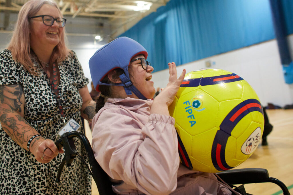 A girl in a wheelchair is being pushed around an indoor court, she has a ball in her hands