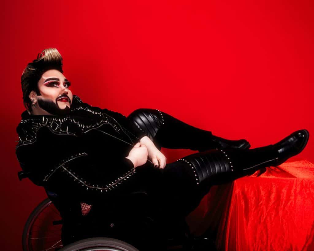 Crip Ladywood, a white person dressed in studded leather with coiffed blonde hair and full drag make-up, reclines in a wheelchair against a red backdrop.