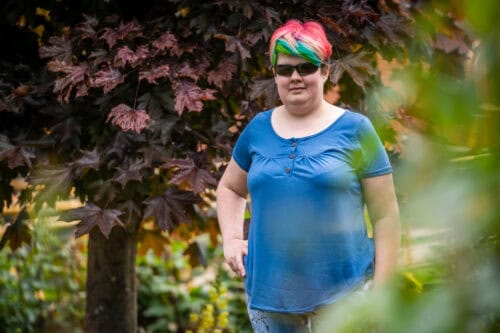 Lucy, a woman wearing sunglasses with short, rainbow-coloured hair, standing among green foliage.