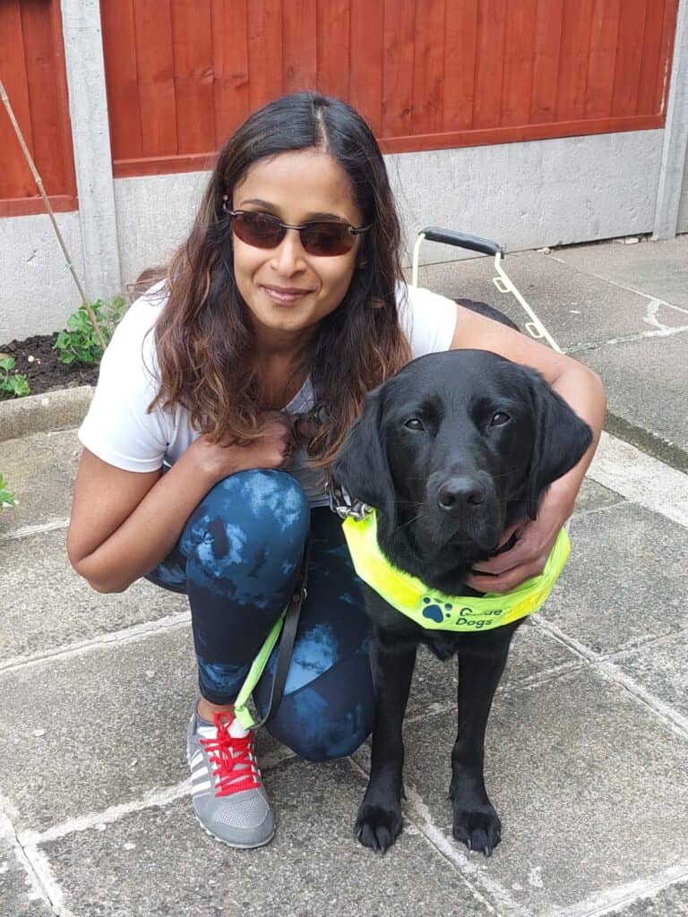 Jaina Mistry, an Asian woman wearing sunglasses, crouching beside her assistance dog Kath.