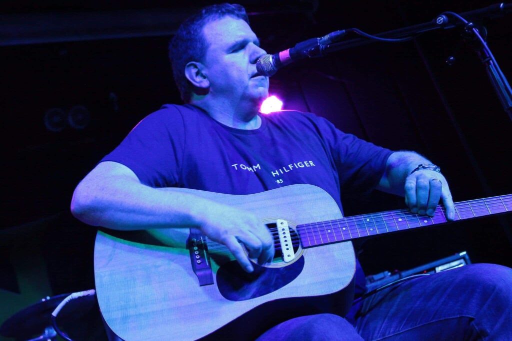 Joe Kenny, a white man sitting playing a guitar and singing into a microphone under blue lighting.