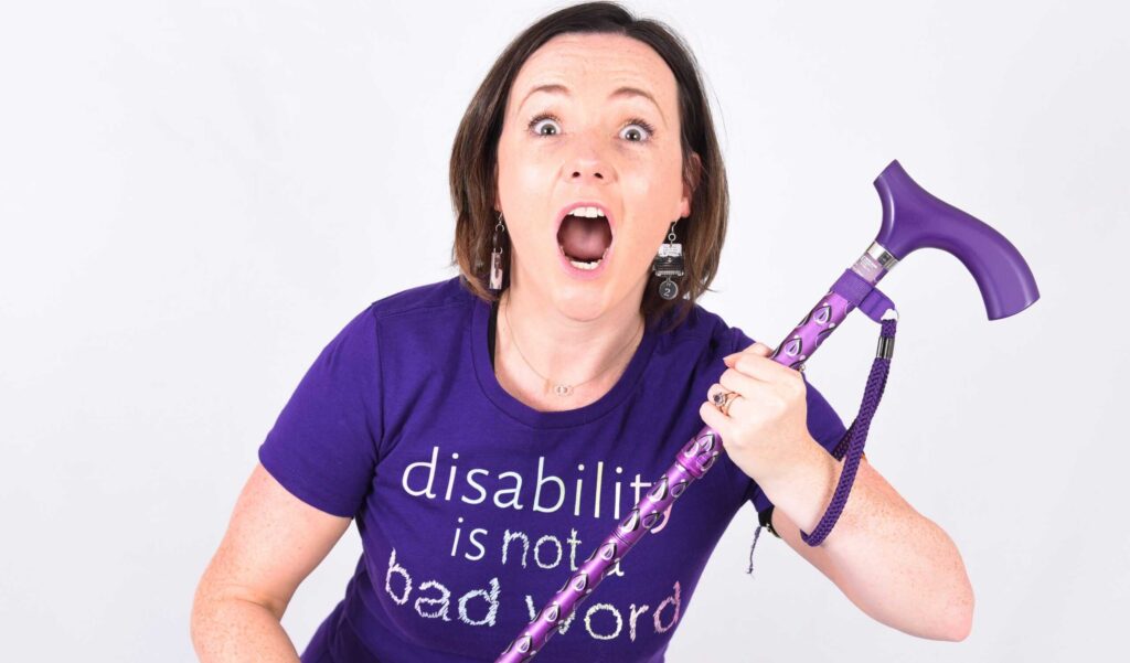Lisette Auton photographed with her mouth open ready to shout. She is holding a purple cane and wearing a purple t-shirt reading 'disability is not a bad word'.