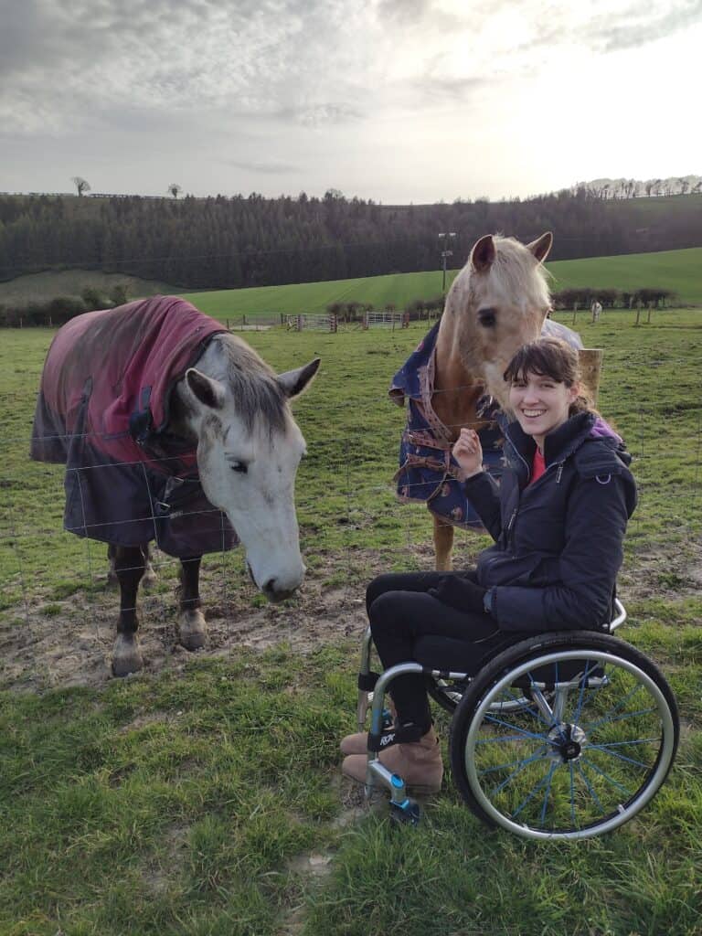 A young woman is petting two horses from her wheelchair, reaching over a fence in a large field.