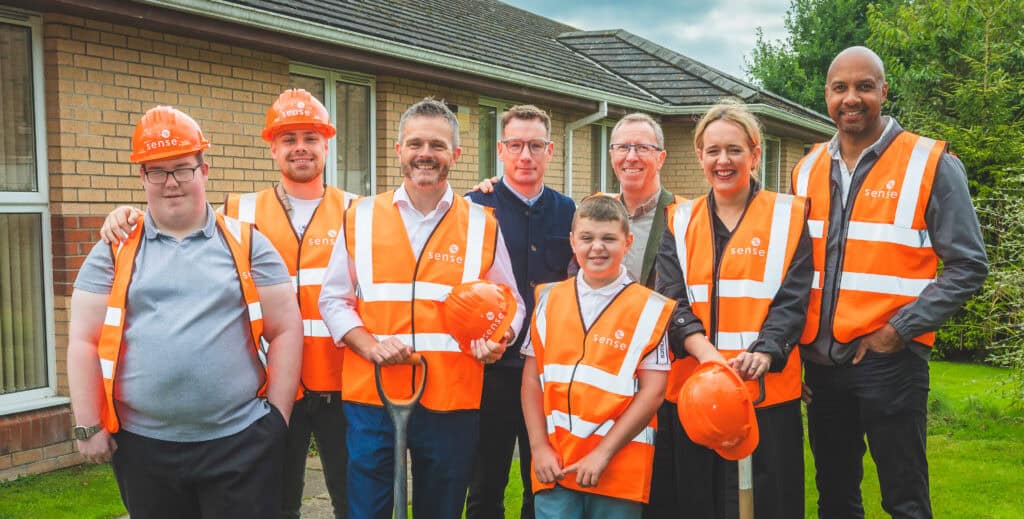 A group of 7 adults and one young boy standing together in a group outside a brick building. They are all wearing orange Sense branded hi vis vests, some of them are wearing orange Sense branded hard hats. A man are the front holds a space.