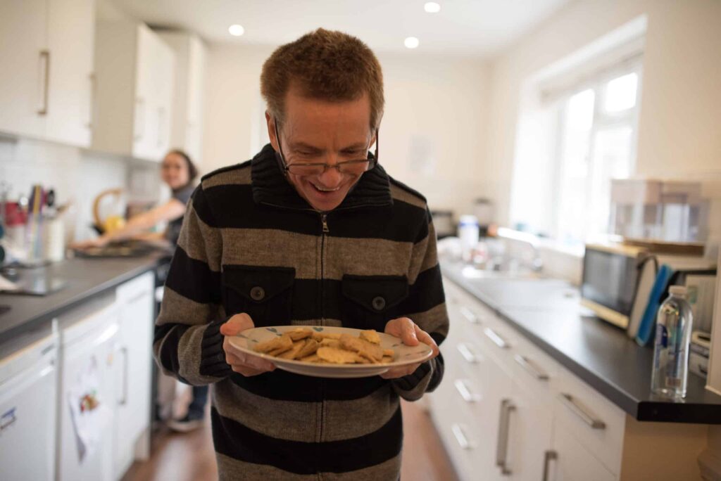 A man walks away from a kitchen, he's smiling as he looks down at a plate of biscuits