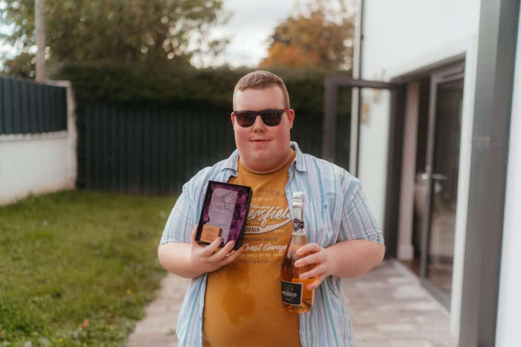 Jamie is wearing sunglasses and standing outside. He's holding his Sense Award and a bottle of fizz.
